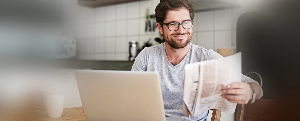 Mann liest Zeitung und sitzt am Laptop