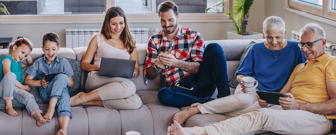 Mehrgenerationenfamilie sitzt lachend auf dem Sofa, nutzt gemeinsam Laptop, Smartphone und Tablets in gemütlicher Wohnzimmeratmosphäre.